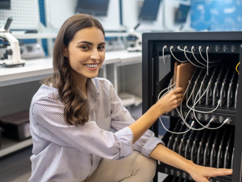 Equipment. Young adult woman crouching near open dedicated charger box smiling at camera touching charging board indoors